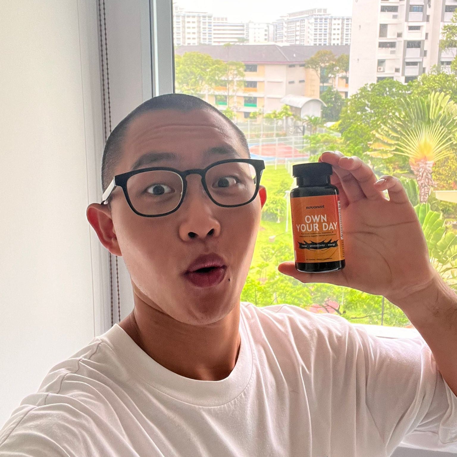 Person holding a supplement bottle in front of a window with a view of buildings and greenery.